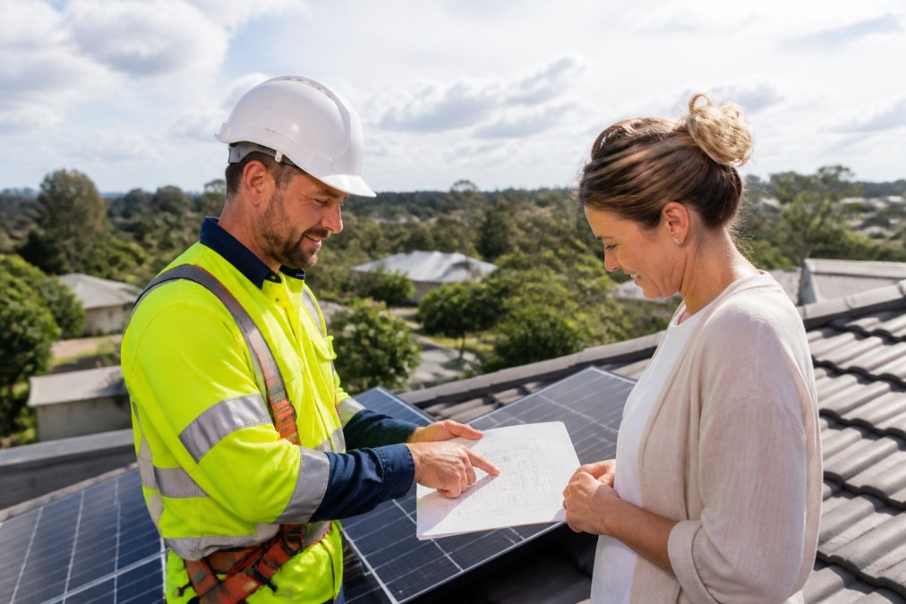 CEC accredited solar installer in high-visibility safety gear and hard hat discussing system design specifications with homeowner on residential rooftop during daylight, with solar panels visible in background