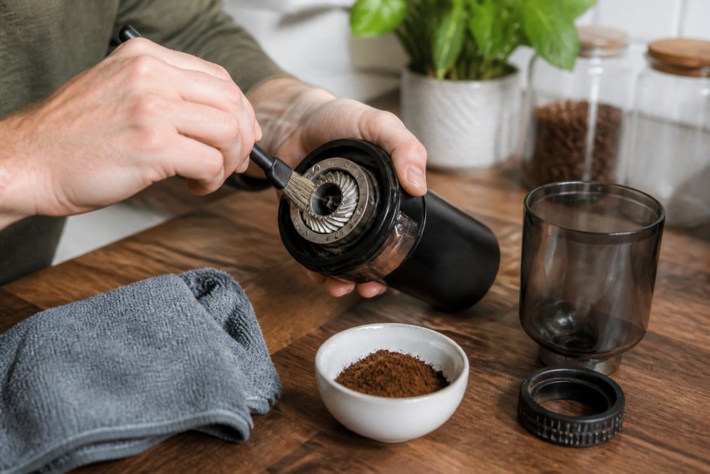 Hands using a stiff brush to clean coffee grounds from the conical burrs of a disassembled burr coffee grinder.