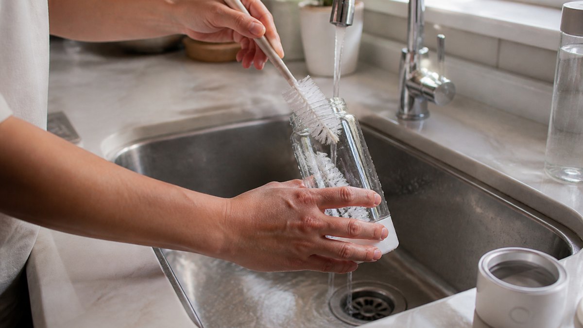 Close-up of hands using a soft brush to clean the interior of a hydrogen water bottle under warm running water at a kitchen sink