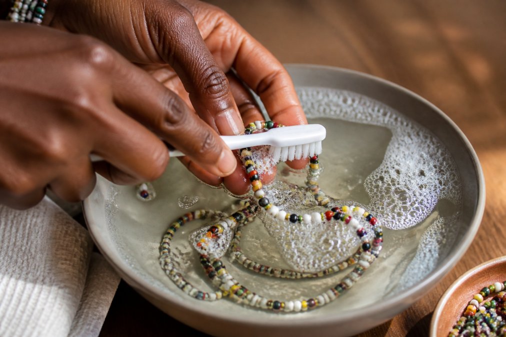 Close-up of hands gently cleaning beaded jewelry with a soft-bristle toothbrush in a shallow bowl of soapy water, showing delicate cleaning technique with natural afternoon lighting
