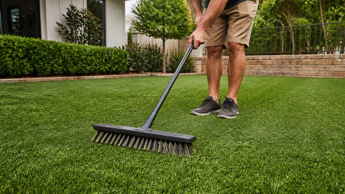 Homeowner using a stiff-bristled turf brush on residential artificial grass in backyard, showing proper brushing technique with fibers standing upright after brushing