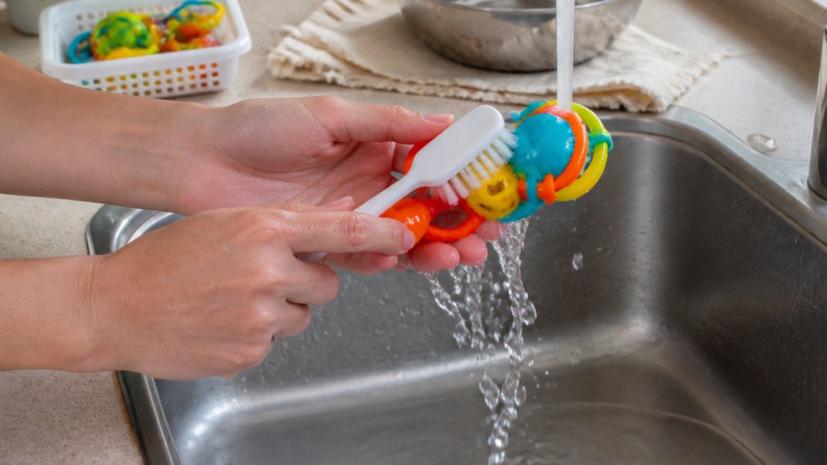 Close-up of hands scrubbing a colorful plastic bird toy with a soft brush under warm running water in a sink