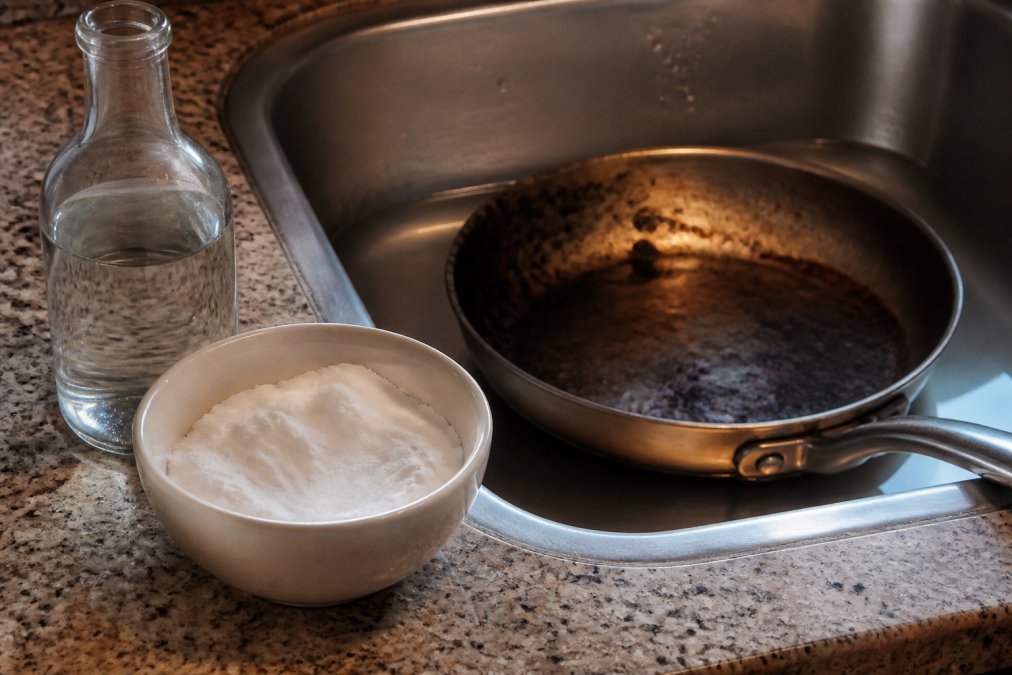 Close-up of a kitchen counter with a small white bowl filled with thick baking soda paste, a bottle of white vinegar beside it, and a darkened stainless steel pan soaking in a stainless sink under warm overhead lighting