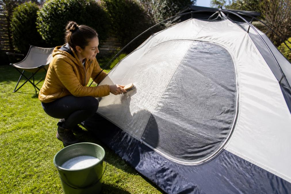 A person in outdoor clothing gently scrubbing the fabric of a camping tent with a soft brush and bucket of soapy water, set up in a grassy backyard on a sunny day