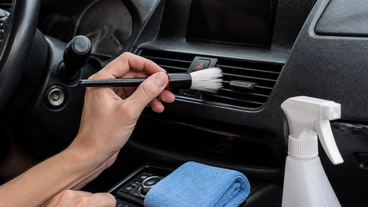 Close-up of a person's hands using a soft detailing brush to clean the narrow slats of a car air vent on a dashboard, with a microfiber cloth and spray bottle visible nearby