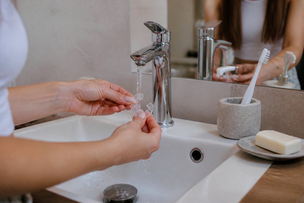 Close-up of hands rinsing clear aligners under lukewarm running water in a bathroom sink with a soft-bristled toothbrush and unscented soap nearby on the counter