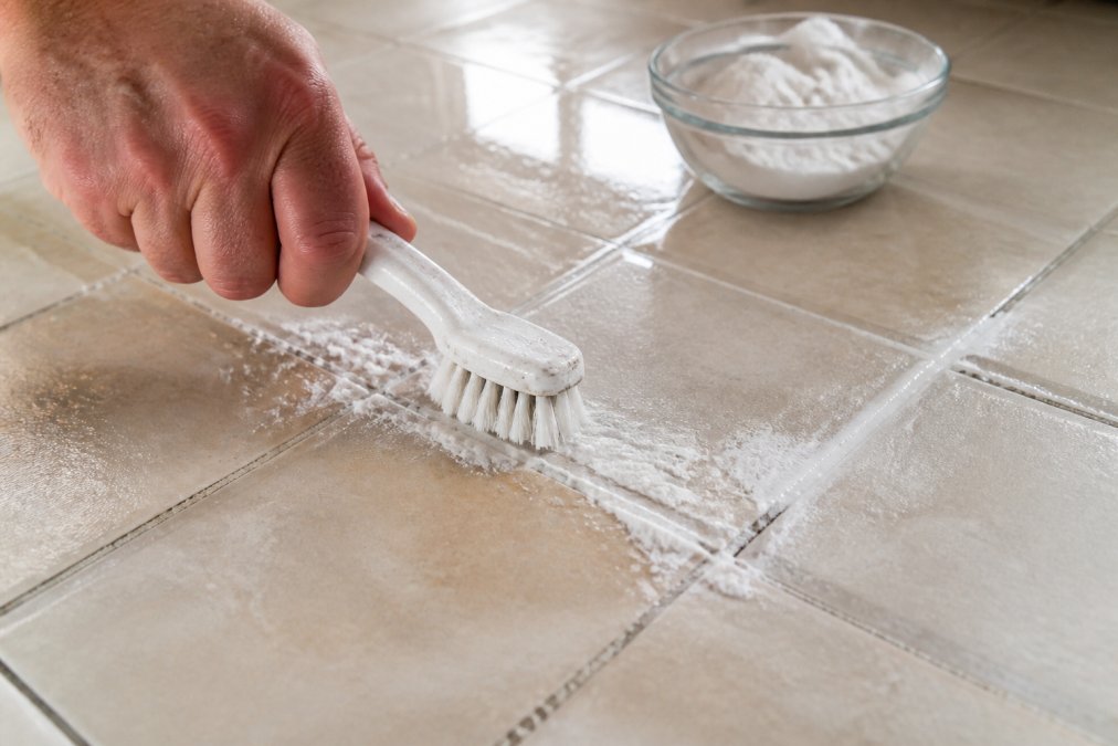 Stiff-bristled brush scrubbing baking soda paste to clean deeply stained grout on a ceramic tile floor