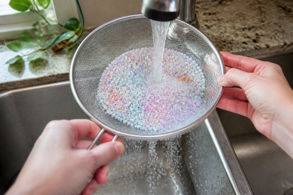 Close-up of hands rinsing acrylic beads in a mesh strainer under warm running water with visible dish soap bubbles and water droplets, set in a bright kitchen sink with natural daylight
