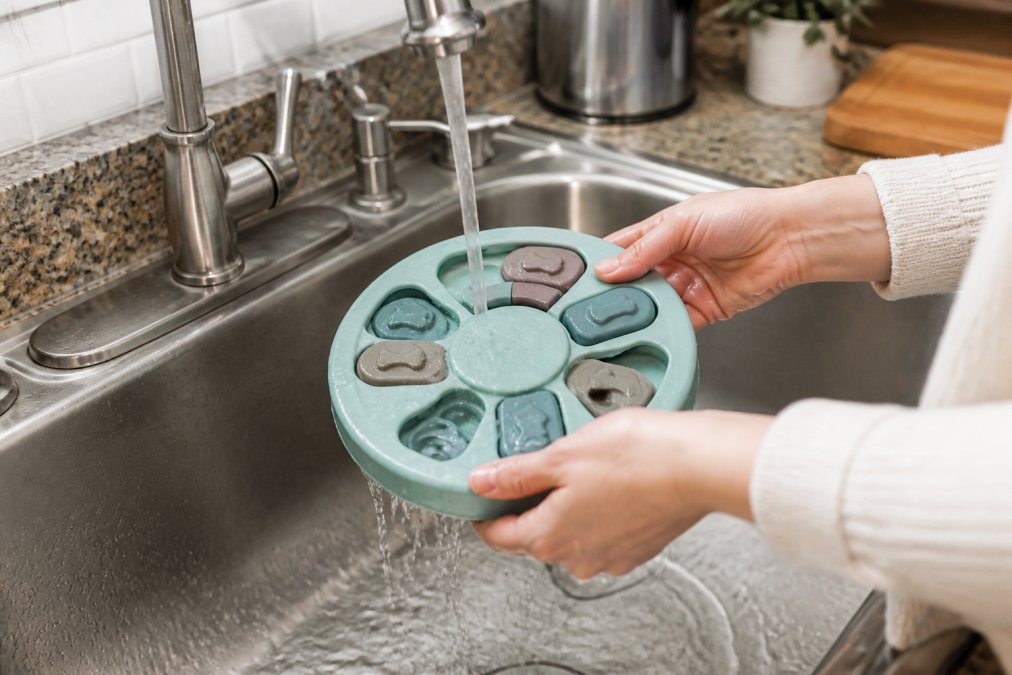 Close-up of hands rinsing a puzzle feeder under warm running water in a kitchen sink, showing water flowing over the toy's compartments and sliding covers