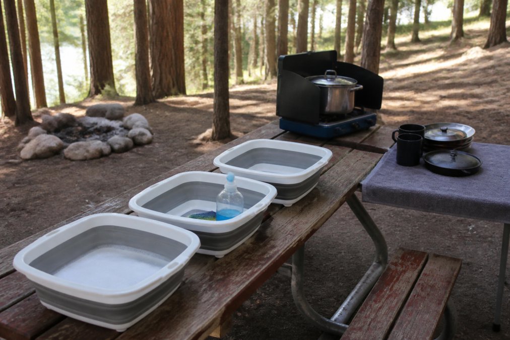 A campsite dishwashing station with three collapsible basins on a picnic table, a camp stove heating water, and cookware drying on a towel in a forest setting