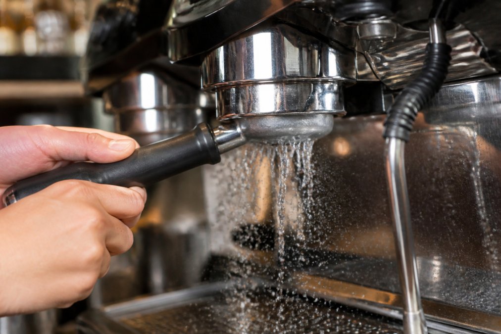 Close-up of hands performing backflushing on espresso machine group head, water spraying from portafilter, blind basket inserted, stainless steel group head visible