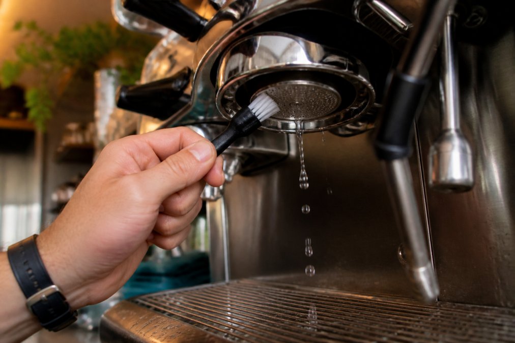 Barista's hands holding a soft nylon group head brush, gently scrubbing the shower screen of an espresso machine group head, water droplets visible, professional espresso machine in focus