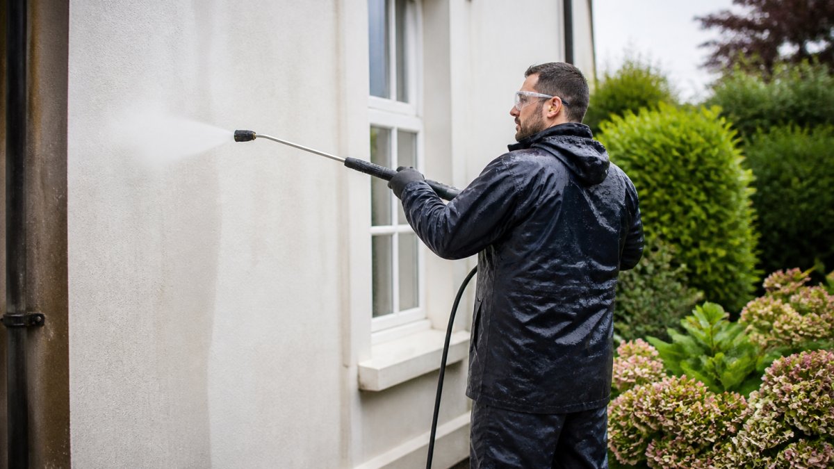A professional exterior cleaner in protective gear using a low-pressure soft wash lance on a rendered house wall, with a garden and established plants visible nearby, demonstrating careful surface-specific cleaning