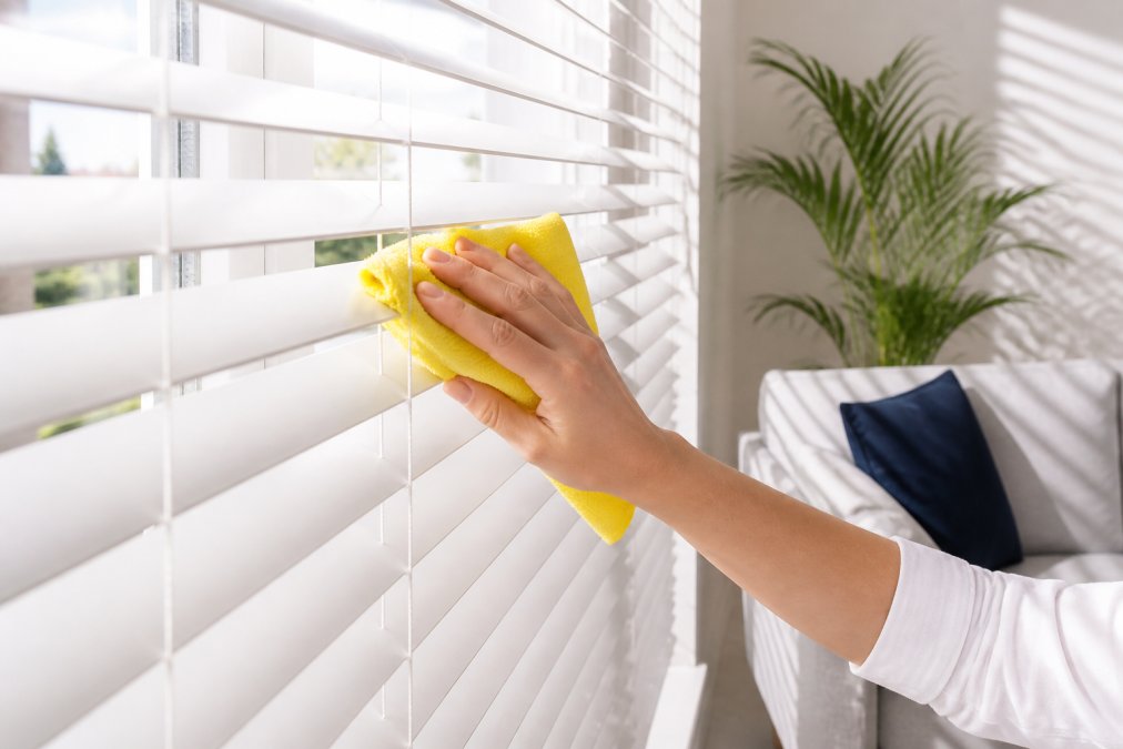 A close-up of a person using a yellow microfiber cloth to dust the horizontal slats of white faux wood Venetian blinds in a bright living room, with sunlight streaming through the window