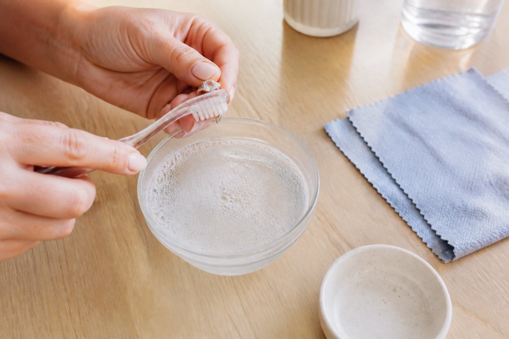 Close-up of hands gently cleaning a gemstone ring with a soft-bristled toothbrush over a bowl of warm soapy water, lint-free cloth nearby on a light wooden surface