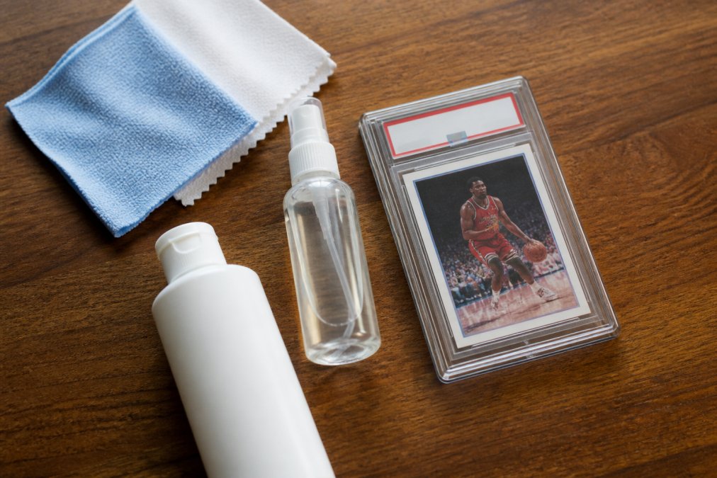 A flat lay of cleaning supplies on a wooden table: microfiber cloths, a small spray bottle of distilled water, a bottle of plastic polish, and a PSA graded card slab in a protective case.