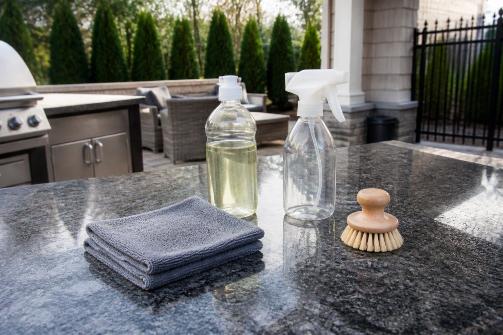 Homeowner arranging cleaning supplies on countertop including microfiber cloths, pH-neutral cleaner bottle, spray bottle, and soft-bristled brush in natural daylight