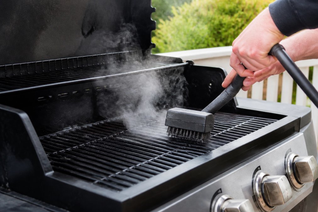 Hands holding a steam grill brush over hot grates with steam rising visibly, showing the brush actively cleaning carbonized buildup in a backyard setting with natural daylight