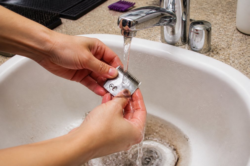 Close-up of hands holding metal clipper blades under warm running water, with hair and debris being rinsed away into a white sink
