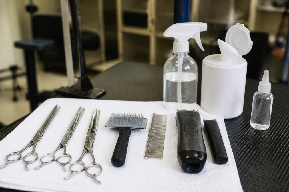 Professional grooming tools arranged neatly on a clean white towel next to bottles of disinfectant spray, wipes, and clipper oil in organized workspace