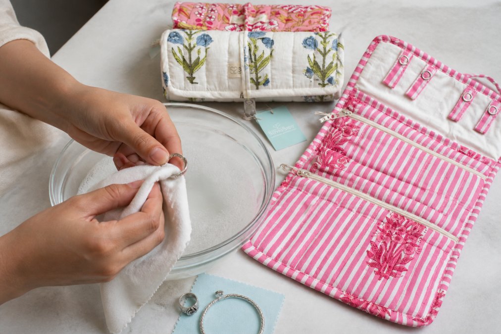 Close-up of hands gently cleaning a delicate silver ring with a soft microfibre cloth over a bowl of soapy water in natural daylight