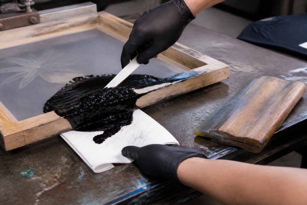 Screen printer using a plastic spatula to card excess ink from a wooden frame onto a towel