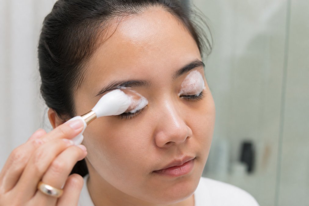 Woman gently cleaning her eyelash extensions with a soft foam cleanser and a small brush in a bright bathroom