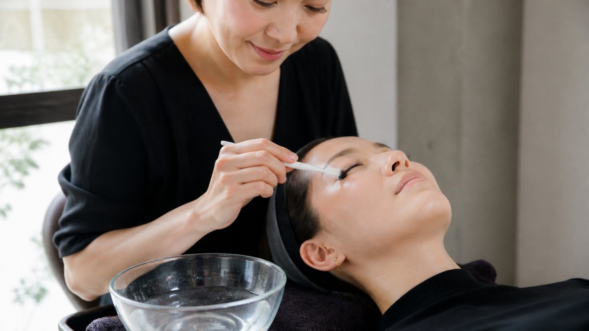 Professional demonstrating proper lash extension cleansing technique with hands gently working a soft brush through extensions over a bowl of lukewarm water in bright natural lighting