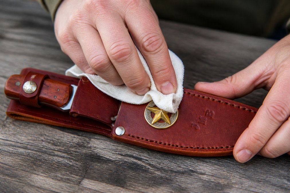 Close-up of hands gently wiping a leather knife sheath with a damp cloth, showing proper technique and care during cleaning