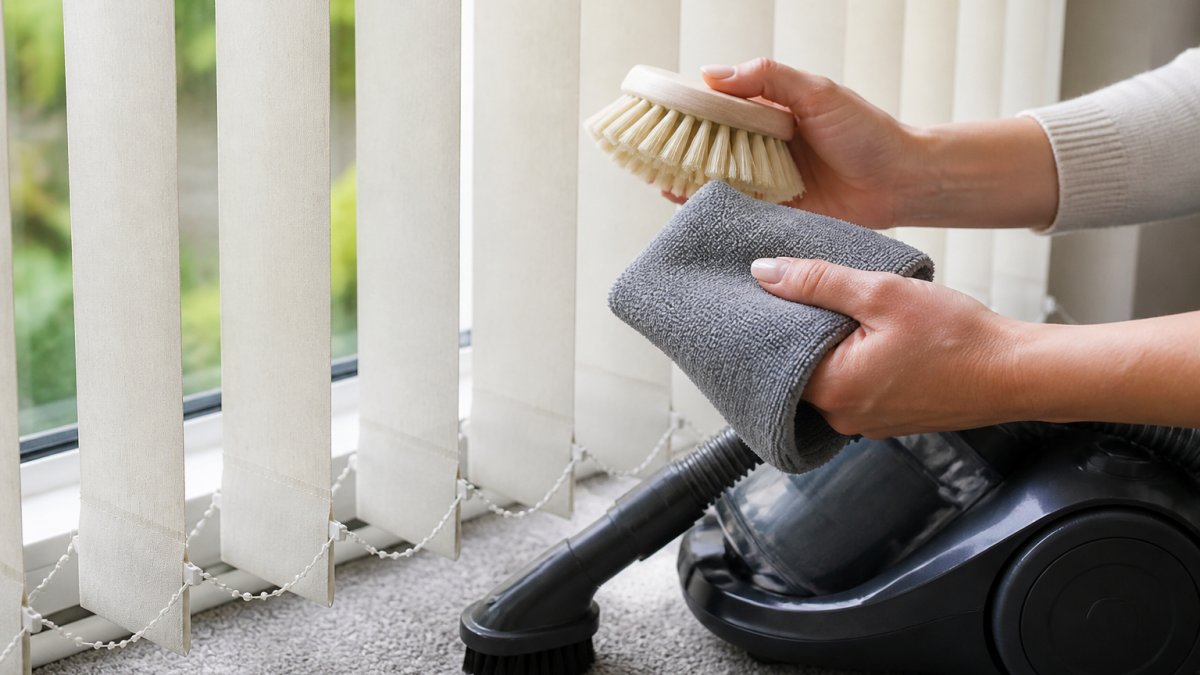 Close-up of hands holding soft-bristled brush and microfibre cloth next to vacuum cleaner with brush attachment, positioned near vertical blind slats in natural daylight