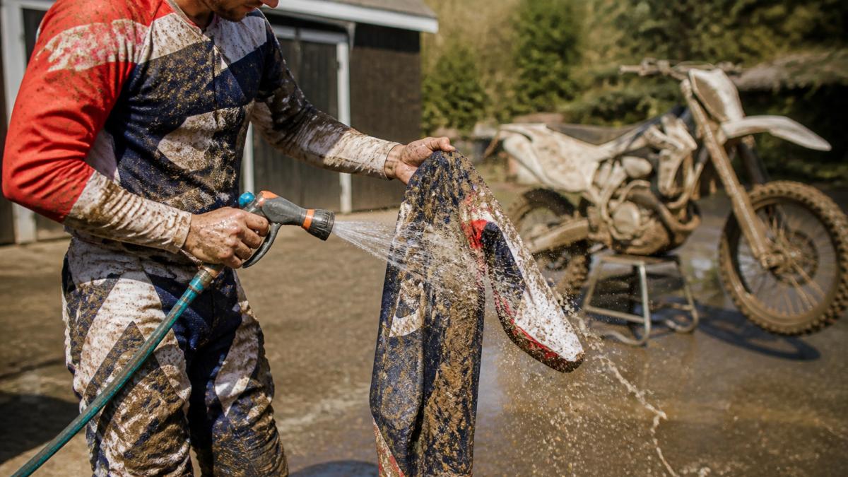 Rider in muddy motocross jersey and pants rinsing gear with a garden hose after a ride, water spraying off caked-on mud in bright outdoor sunlight