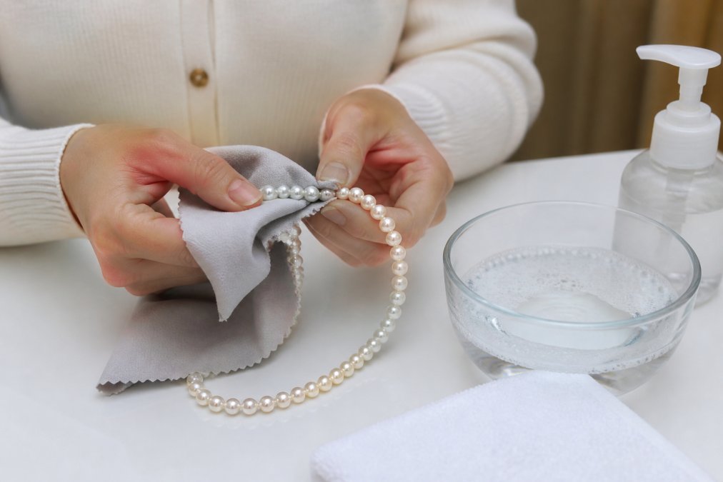 Close-up of hands gently wiping a natural pearl necklace with a soft microfiber cloth on a clean white surface, with a small bowl of lukewarm water and mild soap nearby