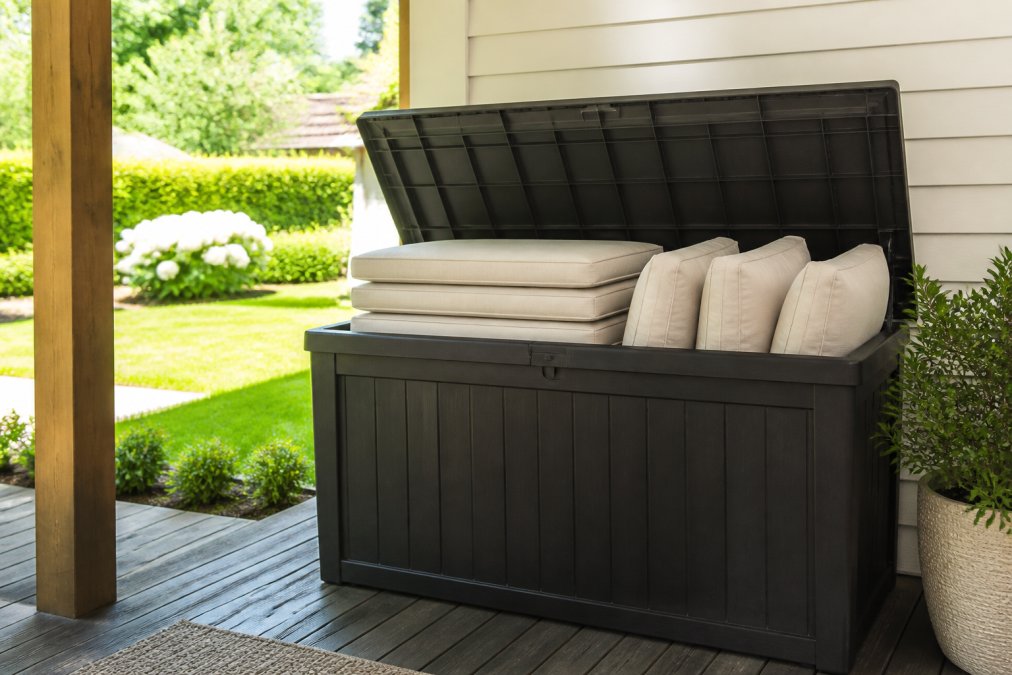 Neatly stacked clean outdoor cushions stored in a large weather-resistant storage bin on a covered patio, with a tidy garden visible in the background