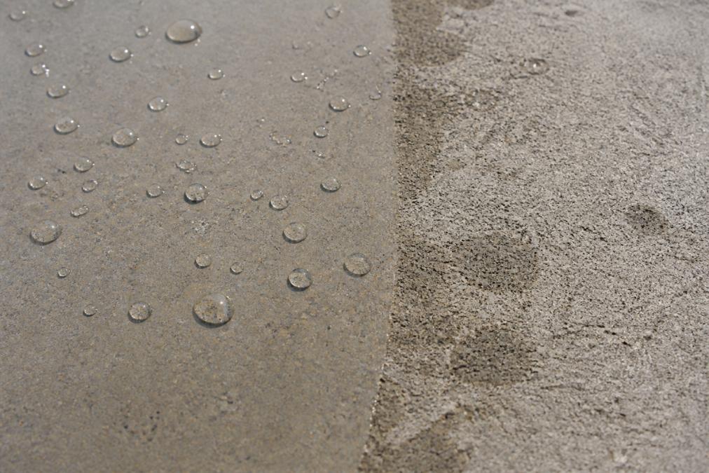 Close-up of water droplets beading on sealed concrete surface in bright daylight, contrasted with water absorption into unsealed porous concrete beside it, showing clear surface tension difference
