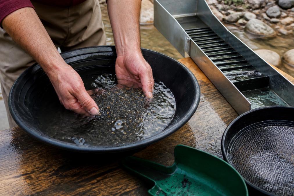 Close-up of hands in a plastic pan with water, gently swirling raw placer gold flakes and black sand concentrates; small sluice box and wet screening tools visible on a wooden table outdoors, natural daylight