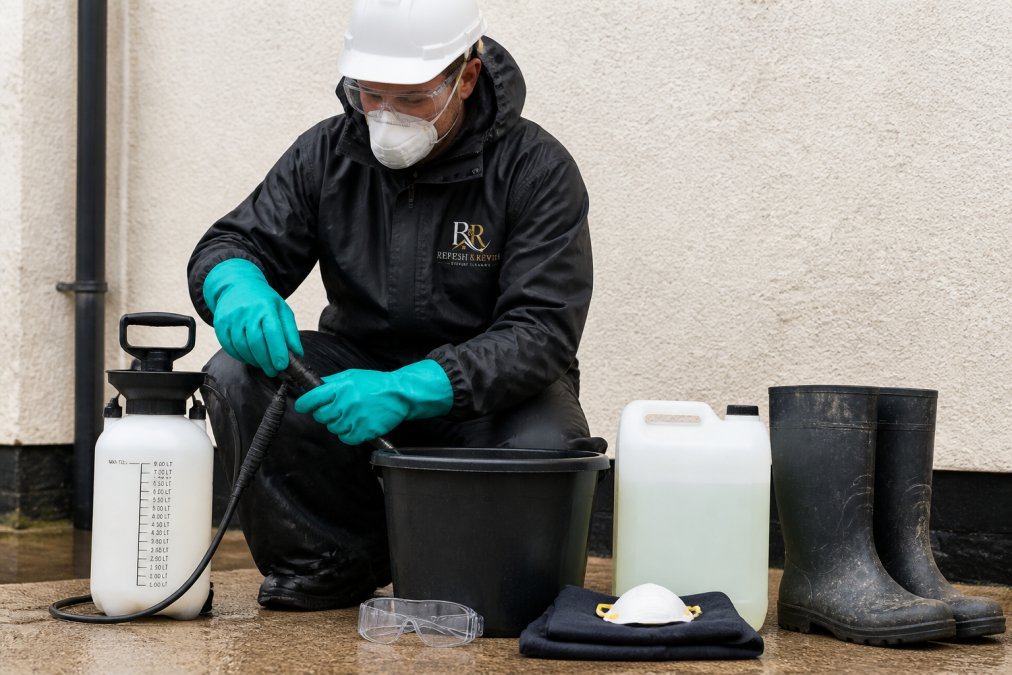 Worker wearing full PPE including safety goggles, chemical-resistant gloves, protective clothing, and hard hat while preparing cleaning equipment and supplies on ground level