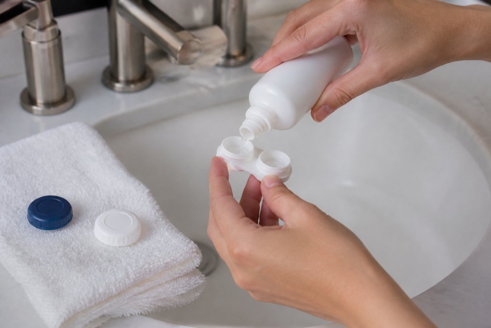 Close-up of hands cleaning a contact lens case with solution at a bathroom sink, with a bottle of multi-purpose solution and clean towel nearby