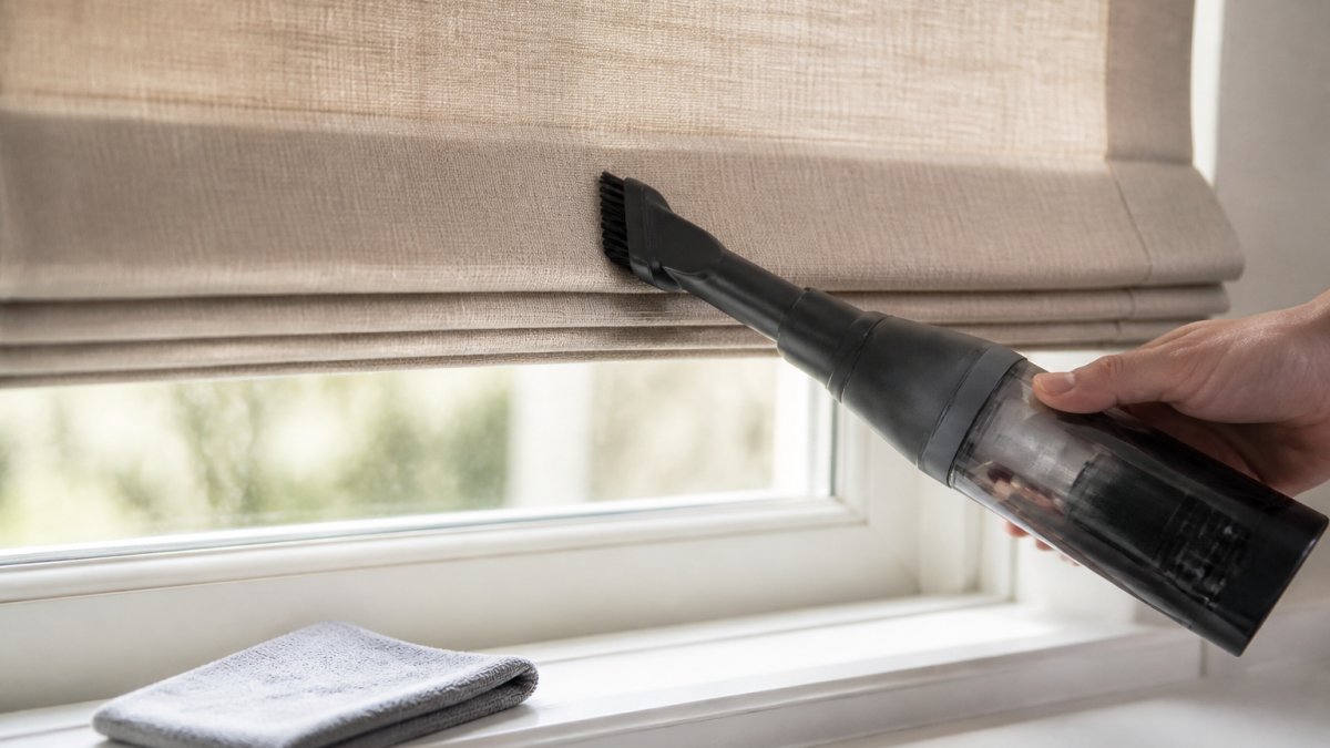 Hand using a soft vacuum brush to gently clean roman shades on a window frame