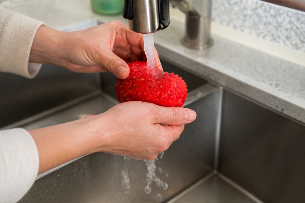 Close-up of hands rinsing a rubber dog toy under warm running water, showing water flowing over the toy's textured surface with visible debris washing away