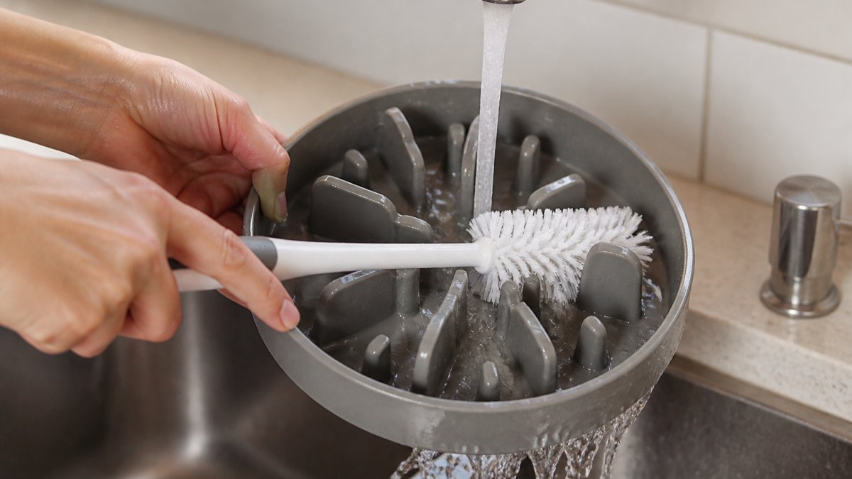 Close-up of hands using a dedicated bottle brush to scrub the grooves and ridges of a slow feeder bowl under warm running water in a bright kitchen sink