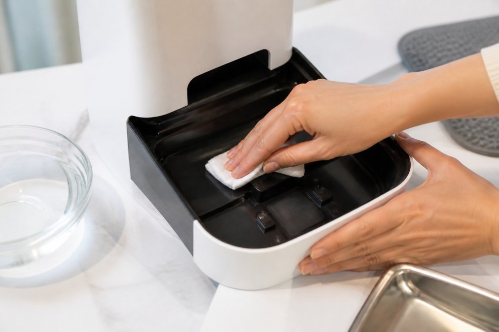 Close-up of hands using a lint-free cloth to gently clean the sensor area inside an automatic pet feeder's base unit with warm water nearby
