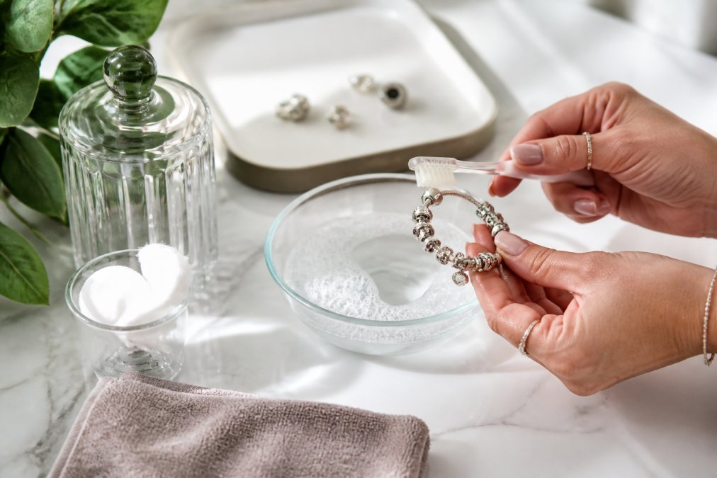 Close-up of hands gently cleaning a sterling silver charm bracelet with a soft-bristled toothbrush in a small bowl of soapy lukewarm water on a white marble surface, soft natural window light illuminating the silver details