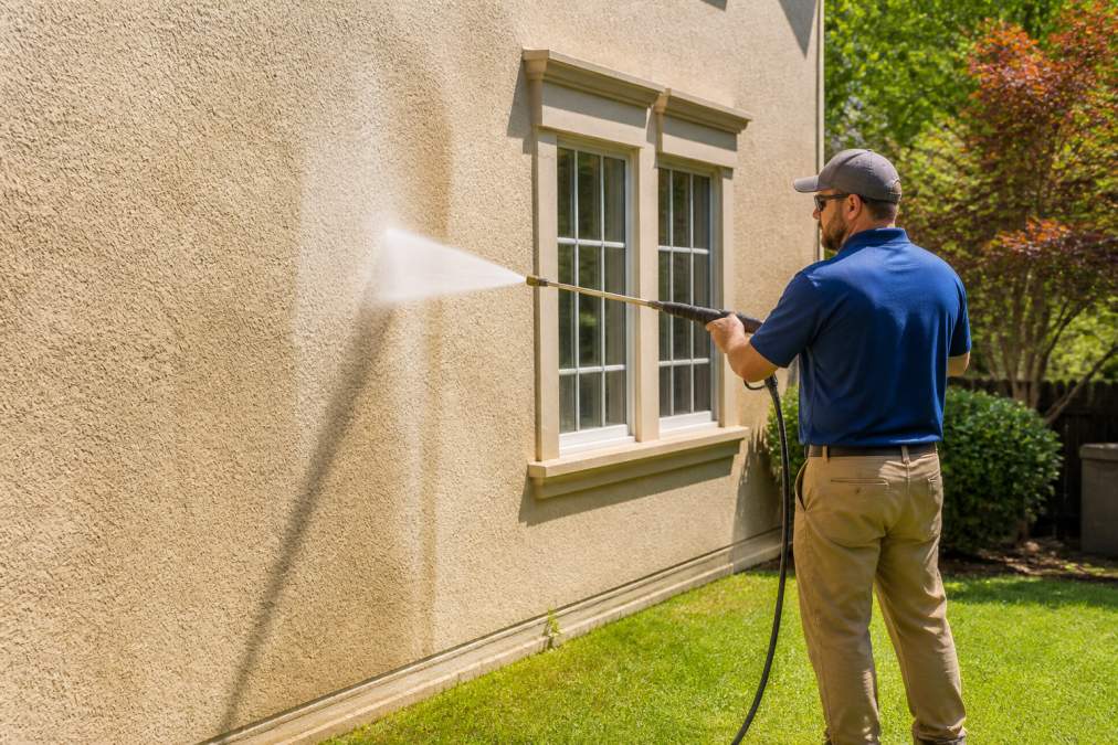 A homeowner in work clothes using a pressure washer with a wide fan spray nozzle on a beige stucco house exterior, standing several feet back from the wall on a sunny day