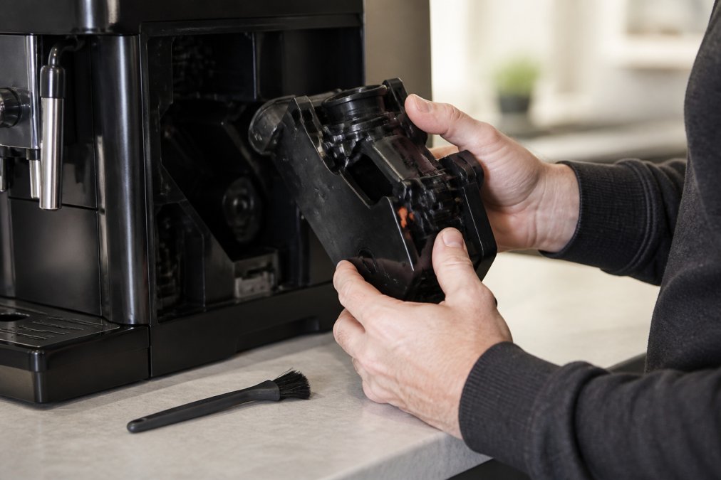 Close-up of hands removing the brew group from a super automatic espresso machine, showing the part detached on a clean kitchen counter with a soft brush nearby