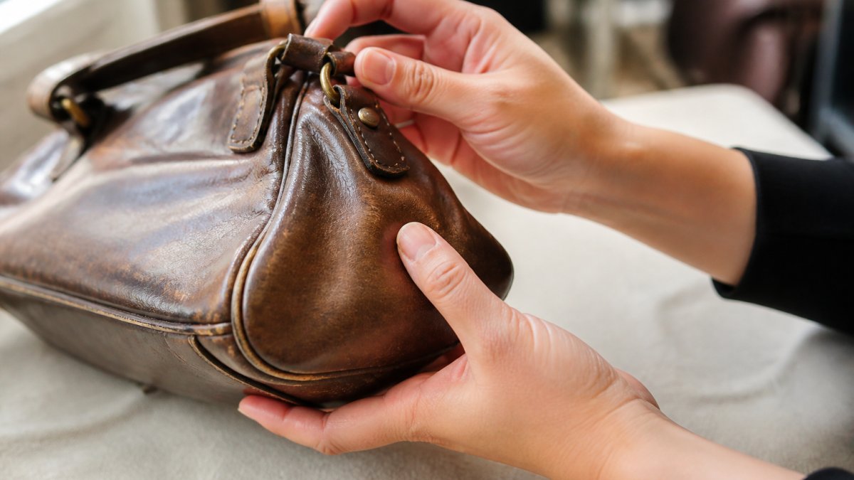 Close-up of hands carefully examining a vintage leather handbag under natural light, showing detailed inspection of leather surface texture, seams, and metal hardware for condition assessment