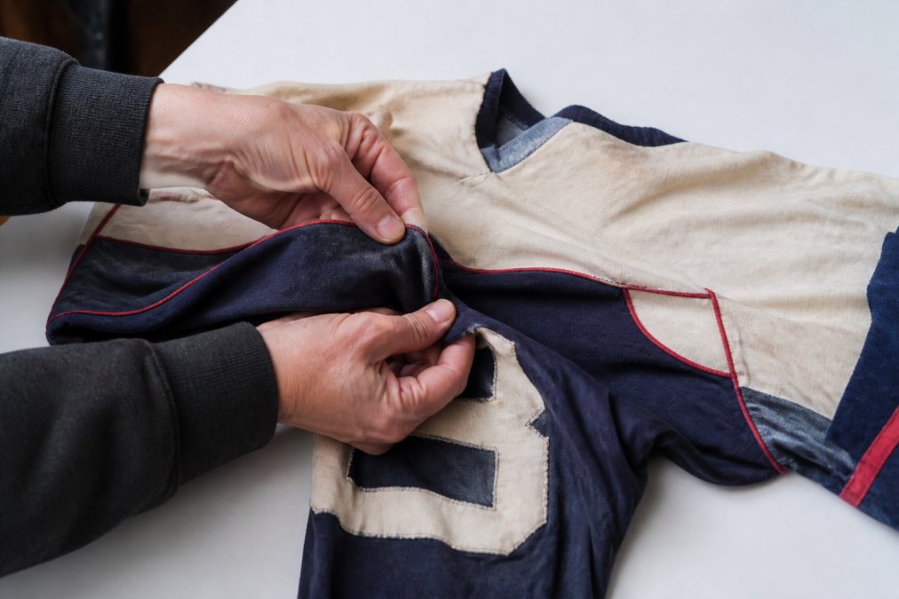 Hands carefully inspecting a vintage sports jersey laid flat on a clean white surface, examining seams and fabric condition under natural daylight