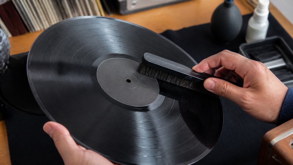Hands holding a vintage vinyl record while using an anti-static brush to gently remove dust from the grooves.