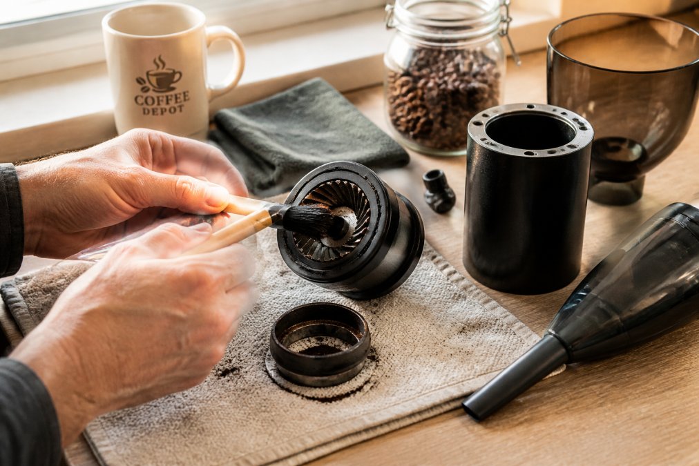 Close-up of hands using a small stiff-bristle brush to sweep coffee grounds from the burrs of a disassembled burr grinder on a kitchen counter, with a microfiber cloth and small vacuum nearby, warm morning light