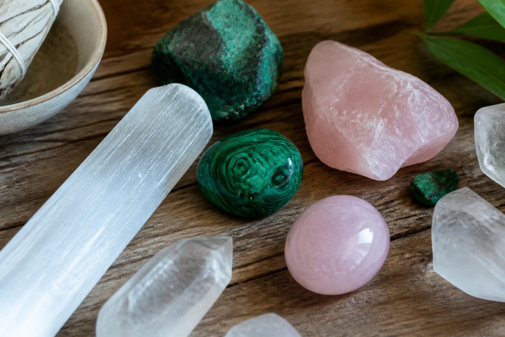 Close-up flat lay of several raw and polished crystals, selenite, rose quartz, and malachite, arranged on a worn wooden surface with soft, diffused natural window light