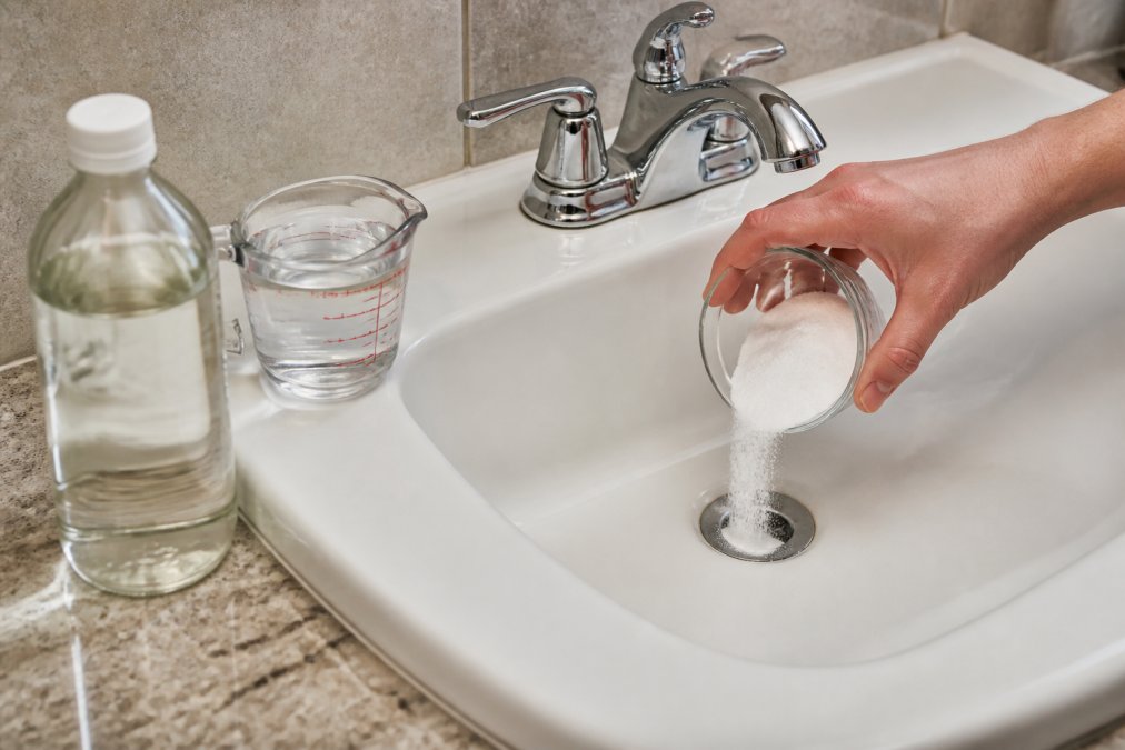 Close-up of a hand pouring baking soda into a white bathroom sink drain, with a bottle of white vinegar and a measuring cup visible on the counter beside the sink under warm bathroom lighting